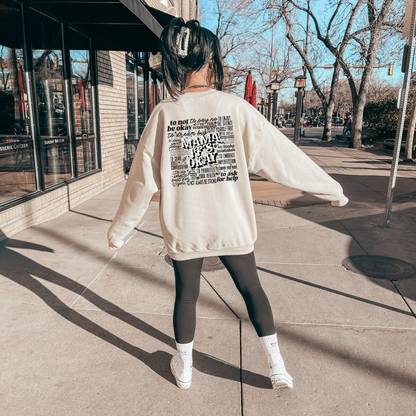 A person standing on a sidewalk, facing away from the camera, wearing a beige crewneck sweatshirt with text on it, paired with dark leggings and white shoes.